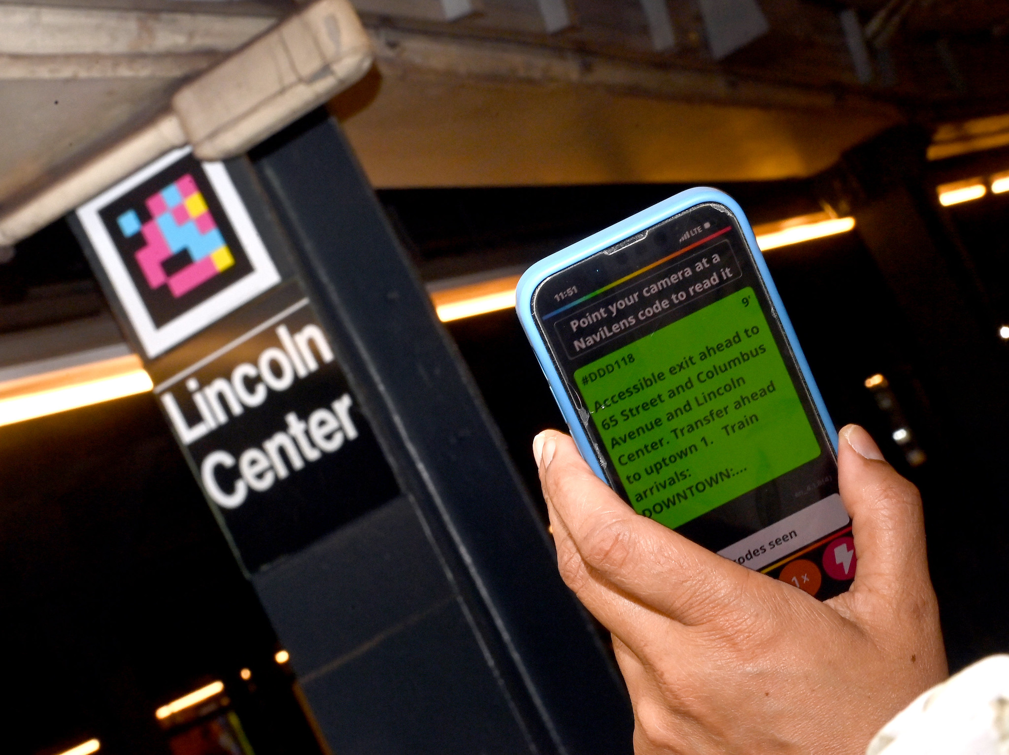 Cell phone shows Navilens wayfinding instructions with accessibility QR code visible in background above Lincoln Center sign.