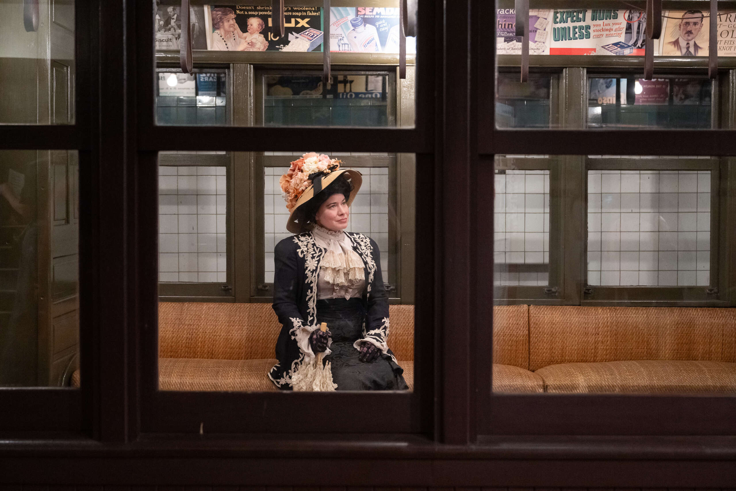A woman dressed in early 1900s clothing sits in a vintage elevated train car.