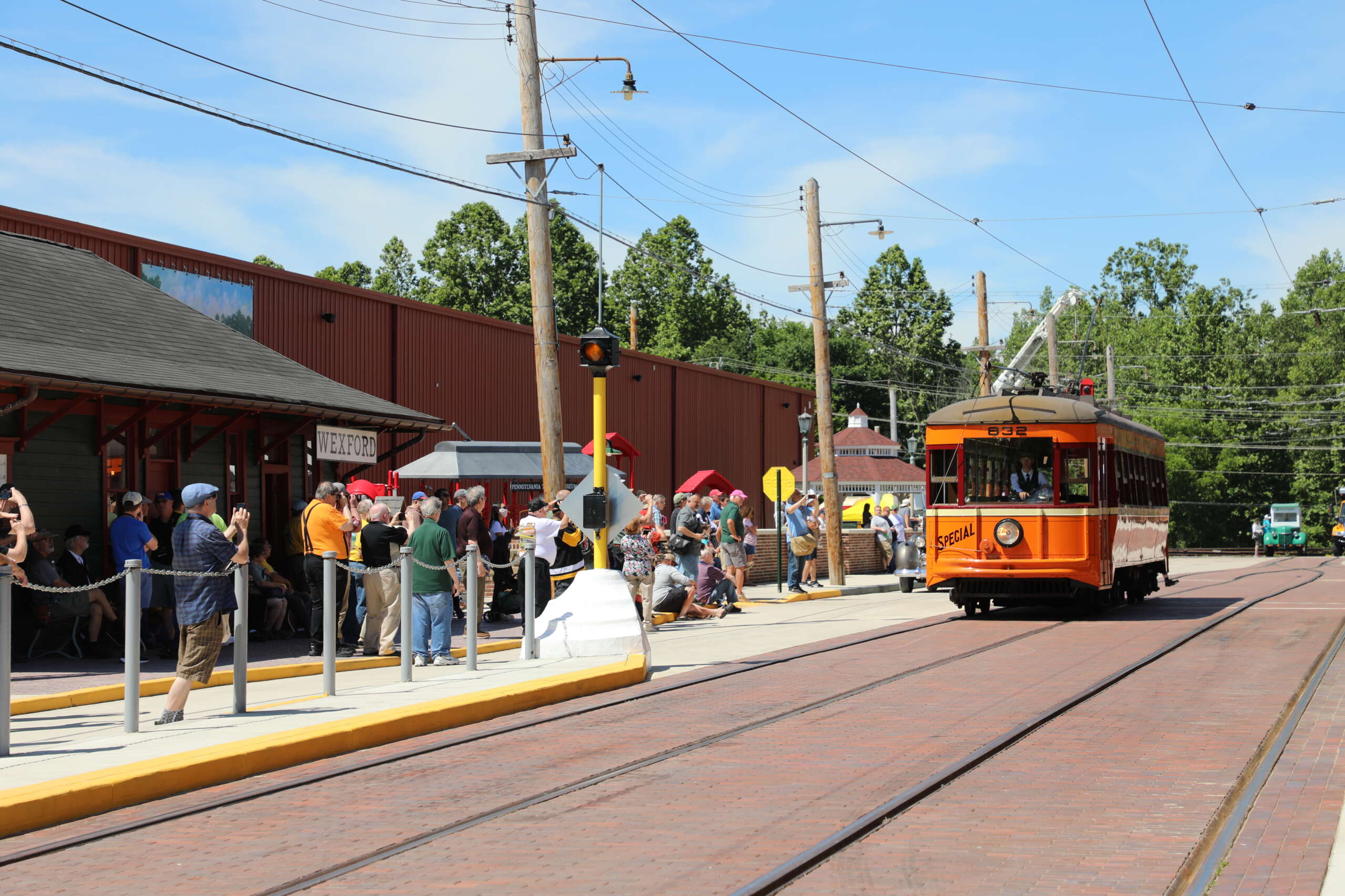 Orange trolley approaches station, with people waiting and taking photographs.