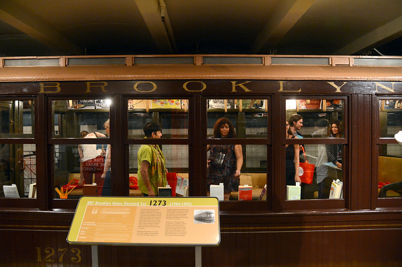 Museum guests are walking through a vintage train on the Transit Museum platform. Train information is posted in front of the train.